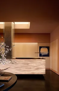 Modern reception area featuring a glowing backlit onyx desk, warm terracotta and beige walls, minimalist brass lamp, and abstract art.