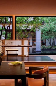 Modern Japanese-style tea room interior featuring a dark walnut armchair with orange leather seat, a low table with books and teacup, and a view of a lush green courtyard with trees and a water feature.