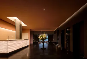 Modern luxury hotel lobby featuring a long illuminated onyx reception desk, dark stone flooring, a central round table with large floral arrangement, and warm terracotta ceiling with recessed lighting.