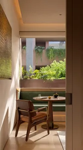 A modern minimalist reading nook featuring a walnut wood armchair with brown leather seat, a round wooden pedestal table, and a built-in green geometric-patterned banquette overlooking a lush vertical garden.