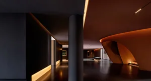 Modern dark hallway featuring a large cylindrical column, continuous LED base lighting, and a dramatic curved orange concrete architectural feature on the right.