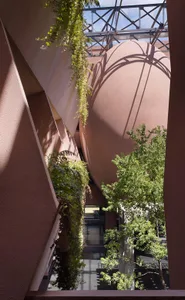 Low-angle view of a modern architectural courtyard featuring a large curved terracotta wall, hanging ferns, and lush green trees under a clear sky with steel truss structure visible above.