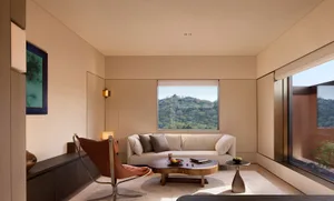 Modern minimalist living room featuring a walnut live-edge coffee table, cognac leather butterfly chair, cream sofa, and large windows overlooking a green mountain with a traditional pavilion.