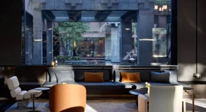 Modern lounge with dark concrete structural beams, large glass walls overlooking a garden, blue-grey sectional sofa with orange accent pillows, and contemporary orange and white lounge chairs.