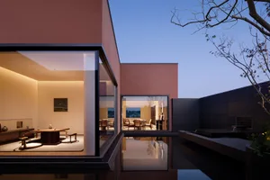 Modern minimalist courtyard house at dusk featuring terracotta plaster walls, a shallow reflective pond, and a warmly lit Japanese-style tea room with low wooden furniture.