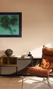 A contemporary living room corner with a brown leather lounge chair, dark wood and brass console, ceramic vase, black sculptural object, and a large framed turquoise ink painting of pine branches on the wall.