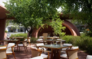 Outdoor terrace featuring wooden tables, white cushioned rattan armchairs, and a large terracotta sculptural arch surrounded by lush green trees and bushes under bright daylight.