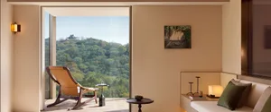 Modern minimalist living room featuring a walnut and tan leather rocking chair, built-in window seat with green velvet cushions, round side tables, and a breathtaking view of a lush green mountain with a traditional Chinese temple.