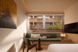 Modern hotel room featuring a green daybed, walnut armchair, round wooden table, large picture window overlooking a pink courtyard with hanging plants, and a textured gold artwork on the wall.