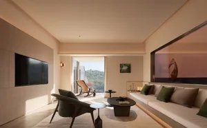 Modern minimalist living room featuring walnut wood armchairs with moss green velvet upholstery, a built-in beige sofa with green and taupe cushions, a black oval coffee table, and large windows overlooking a green hillside.