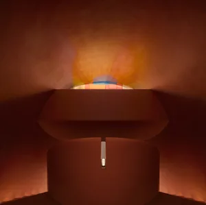 Abstract curved architectural installation in warm terracotta tones with a glowing multicolored light halo emanating from the top center.