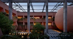View of a brutalist architectural courtyard in dusty pink concrete with large geometric openings, hanging plants, and a massive spherical concrete sculpture at dusk.