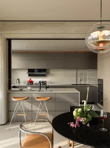 Modern minimalist kitchen with grey matte cabinetry, a central island with two leather bar stools, and a round black marble dining table with a floral arrangement in the foreground.