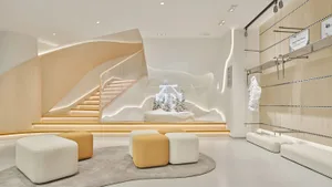 Modern minimalist retail interior with a large illuminated snow globe display, curved oak staircase with integrated lighting, and modular white and mustard ottomans on a grey rug.