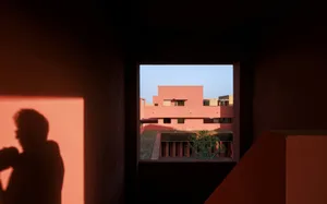 Silhouette of a person looking out from a dark terracotta interior toward a sunlit modernist courtyard with pink and orange buildings and a small tree.