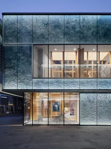 Modern retail building facade featuring large textured blue-green glass panels with an organic wave pattern, revealing a warmly lit minimalist store interior displaying a blue down jacket.