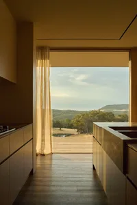 Modern minimalist kitchen with natural oak cabinetry and stone countertops opening to a large glass door with view of forested hills and a pond.