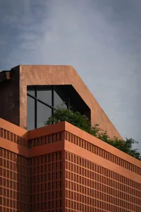 Low-angle view of a modern building featuring a terracotta brick facade with chevron pattern, large black-framed windows, and flowering plants on a concrete ledge against a blue sky.