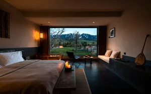 Modern bedroom interior featuring a large bed with taupe linen, a freestanding copper bathtub, built-in window seat, and dramatic mountain landscape visible through floor-to-ceiling windows.