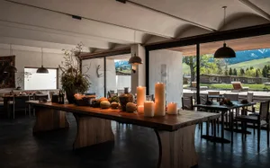 現代阿爾卑斯風餐廳，中央為回收木長餐桌，桌上擺放發光立柱蠟燭、陶器花瓶與乾燥花藝，大片玻璃門外可見山景。