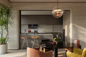 Modern open-plan dining area with round black table, terracotta and mustard velvet chairs, bar stools, and a minimalist taupe kitchen in the background.