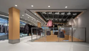 Modern minimalist clothing store interior with large oak columns, white floors, glass railings, and clothing hanging from an overhead industrial grid system under recessed lighting.