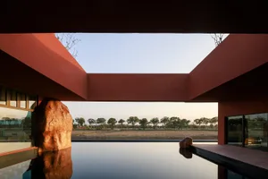Modern rammed earth courtyard house featuring a large reflecting pool, dramatic red earthen walls, and a massive natural boulder under a clear sky.