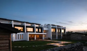 Modern two-story farmhouse with white stucco walls, dark wood accents, and stone masonry base, illuminated at dusk with wooden pathways and stone walls in the garden.