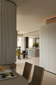 Modern open-plan dining area featuring a prominent curved wood column, neutral wood tones, textured dining chairs, and a view into a minimalist kitchen with a large floral arrangement.
