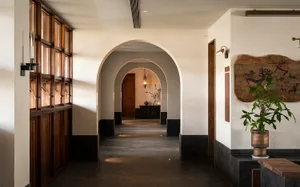 Interior view of a long arched hallway featuring dark wood paneling on the left, textured off-white walls, dark baseboards, a potted plant on a wooden bench, and a large abstract wooden wall art with carved animal motifs on the right.