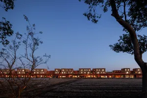 Row of modern terracotta-colored residential buildings at dusk, viewed across a harvested field with foreground trees in Taiwan.