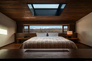 Modern minimalist bedroom with dark wood ceiling, large skylight, striped bedding, and panoramic snow-capped mountain view through wide windows.