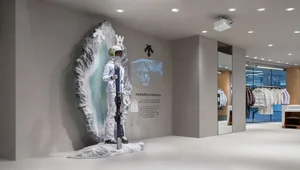 Modern retail space featuring a large textured artificial rock wall with a mannequin dressed in white ski gear holding skis, accompanied by branded graphics and clothing racks in the background.