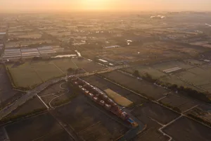 金色時刻航拍畫面，呈現農田、溫室、一座帶有小型游泳池的長型工業或住宅建築群、道路，以及遠方橘色天空下的城市景觀。