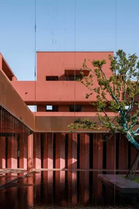 Modern architectural courtyard featuring weathered corten steel walls, a large reflecting pool, and a sculptural tree under a clear blue sky during rainfall.