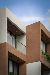Modern two-tone residential building facade featuring red brick on the lower level and off-white concrete upper volume with cantilevered sections and wooden ceiling details under a clear blue sky.