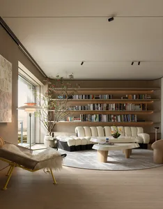 Modern minimalist living room featuring a long curved white modular sofa, irregular oak coffee tables, a woven yellow lounge chair with sheepskin, built-in backlit bookshelves, and a large potted tree under bright natural light from floor-to-ceiling windows.