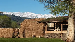 質樸的稻草捆山間小屋，擁有厚實的金黃色稻草牆、木製屋頂結構，背後是壯麗的雪山山脈與晴朗藍天。