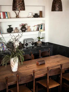 Warm dining room featuring a long reclaimed wood table, dark wooden spindle-back chairs, built-in white shelves with books and ceramics, and a dark plaster lower wall niche.