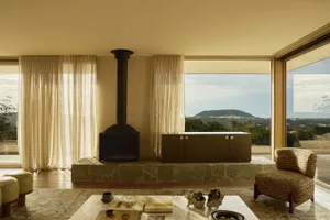 Modern living room featuring a black metal fireplace on a raised stone platform, olive green sideboard, woven armchair, and large windows overlooking rolling hills and mountains.