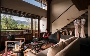 Modern hotel lounge with walnut wood paneling, beige sofa, black leather armchair, round coffee tables on a patterned rug, large glass windows overlooking green mountains, and a woman in traditional attire sitting by the staircase.