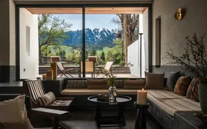 Modern minimalist living room with concrete walls, built-in striped banquette, wooden lounge chair, round coffee table, and large glass opening revealing snow-capped mountains and forest.