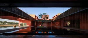 Modern red concrete architectural courtyard featuring a central reflecting pool with a single tree and dramatic linear water curtain in the foreground.