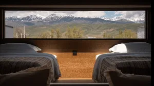 Modern twin hotel room featuring two beds with striped bedding, a full-width wooden headboard, and a large picture window overlooking a dramatic snow-capped mountain range.