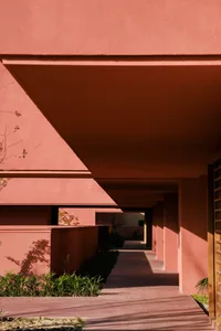 Modern architectural covered walkway with terracotta plaster walls, warm sunlight casting long dramatic shadows on the tiled floor, with green planting along the side.