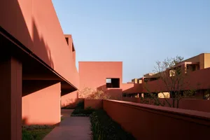 Modern terracotta-colored residential complex with geometric volumes, lush landscaping, and strong afternoon shadows under a clear blue sky.