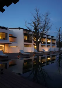 Modern resort pool deck at twilight with white lounge chairs, wooden deck, reflecting pool, and illuminated minimalist white buildings with bare trees against a deep blue sky.