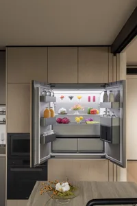 Modern kitchen featuring a large built-in refrigerator with both doors open, displaying fresh fruits, vegetables, and beverages on illuminated shelves, surrounded by light oak cabinetry.