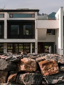 Exterior view of a modern mountain house with white walls, dark wood cladding, large windows, and a foreground dry-stacked stone wall against a backdrop of snow-capped mountains.