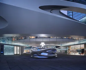 Modern retail atrium featuring a large flowing concrete sculpture on a circular platform, dramatic curved architectural ceiling, and glass storefronts with blue water-like wall panels.